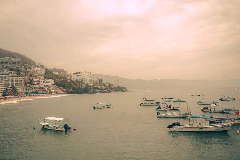 Boats floating near the shore with a view of Puerto Vallarta's coastal buildings under a warm sunset sky.